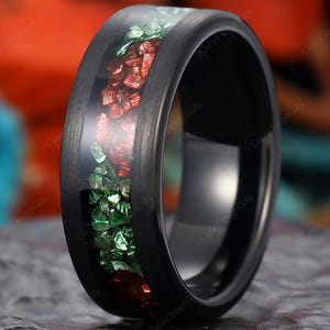 May include: A black tungsten ring with a central inlay of red and green crushed gemstones. The ring has a polished, smooth finish and a dark, modern aesthetic. The gemstones are set in a clear resin, creating a striking contrast.