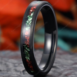 Könnte beinhalten: Ein schwarzer Ring mit einem Band, das mit roten und grünen Fragmenten eingelegt ist. Der Ring ist vor einem verschwommenen Hintergrund mit Orange- und Türkistönen platziert. Der Ring ist eine Nahaufnahme, die die Details des Bandes hervorhebt.