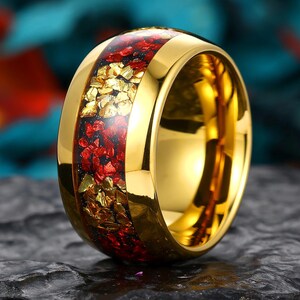 Könnte beinhalten: Ein goldfarbener Ring mit einem breiten, polierten Band und einer mittigen Einlage aus zerkleinerten roten und goldfarbenen Fragmenten. Der Ring hat eine glatte, reflektierende Oberfläche an den Rändern und eine strukturierte, mehrfarbige Einlage.