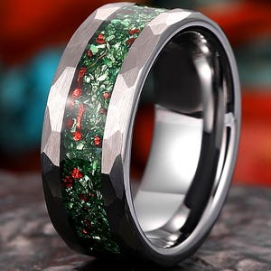 May include: A silver-colored ring with a hammered texture on the outer band. The center of the ring features a green and red stone inlay. The ring is displayed on a dark, textured surface.