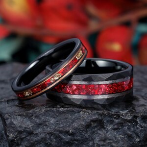 May include: Two black tungsten carbide rings with red and gold inlay. The rings are stacked on top of each other.