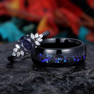 May include: A black and blue wedding ring set with a blue sapphire engagement ring and a matching band. The engagement ring features a blue sapphire stone surrounded by small white diamonds. The band is black with blue glitter.