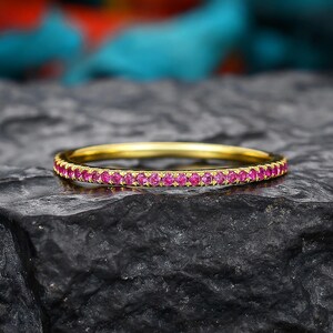 May include: A thin gold band ring featuring a continuous row of small, round, vibrant pink gemstones set along the entire outer circumference. The ring has a polished metallic finish and a uniform, circular design with a slim profile.