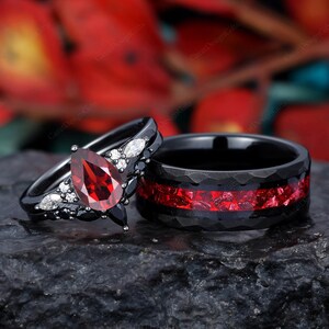 Puede incluir: Un juego de anillos de boda negro y rojo con una piedra preciosa roja en forma de pera y diamantes negros. El anillo de compromiso tiene una banda negra con una fila de diamantes negros y una piedra preciosa roja en forma de pera. La banda de boda es negra con una incrustación roja.