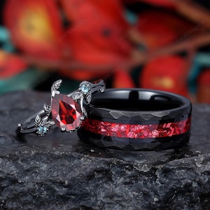 May include: A black and red wedding ring set. The engagement ring features a pear-shaped red gemstone with two smaller blue gemstones on either side. The wedding band is black with a red stripe and a textured finish.