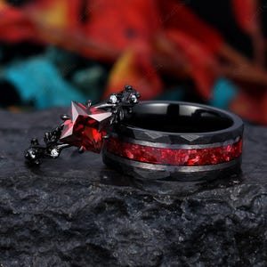 May include: A black and red wedding ring set with a red gemstone. The engagement ring features a star-shaped gemstone with a black floral design. The wedding band is black with a red inlay.