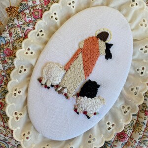 May include: An oval-shaped white fabric piece featuring hand embroidery of a figure in a peach and cream robe with a halo, standing beside two sheep. One sheep is white with black accents, and the other is black with white wool texture.