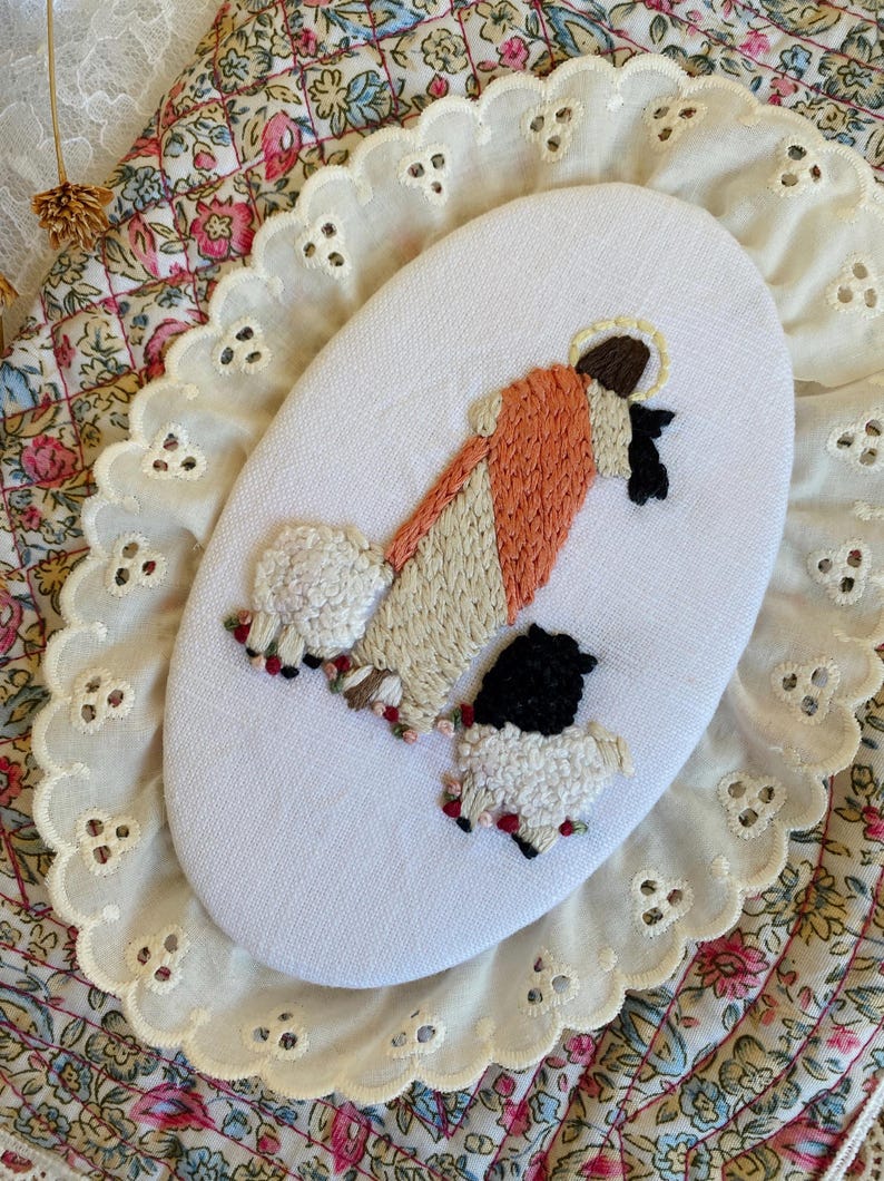 May include: An oval-shaped white fabric piece featuring hand embroidery of a figure in a peach and cream robe with a halo, standing beside two sheep. One sheep is white with black accents, and the other is black with white wool texture.