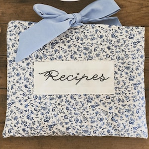 May include: A white fabric recipe holder with a blue floral pattern and a blue ribbon tied in a bow. The holder has a white patch with the word "Recipes" embroidered in black.