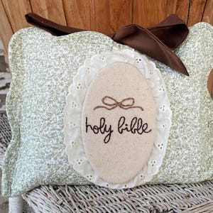 May include: A decorative pillow with a light green floral pattern and a brown bow. An oval-shaped applique with the words "holy bible" embroidered in brown thread is centered on the pillow. The applique has a white lace trim.