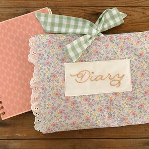 May include: A floral fabric diary cover with a white lace trim and a green gingham bow. The cover has a white patch with the word "Diary" embroidered in brown thread.