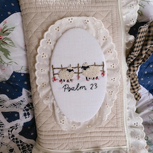 May include: A rectangular beige quilted fabric cover featuring a central oval white embroidered patch. The patch displays two sheep standing by a brown fence with small pink flowers and the black embroidered text Psalm 23. White eyelet lace trim borders the cover and the central oval patch.