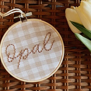 Op de afbeelding: Een bruine en witte gingham stofring met het woord "Opal" geborduurd in bruin garen.