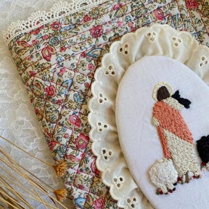 May include: An oval embroidery hoop featuring a stitched figure in a peach robe with a halo, standing beside a white sheep. The hoop is framed by a white eyelet lace trim and rests on a quilted fabric with a floral pattern and lace edge.