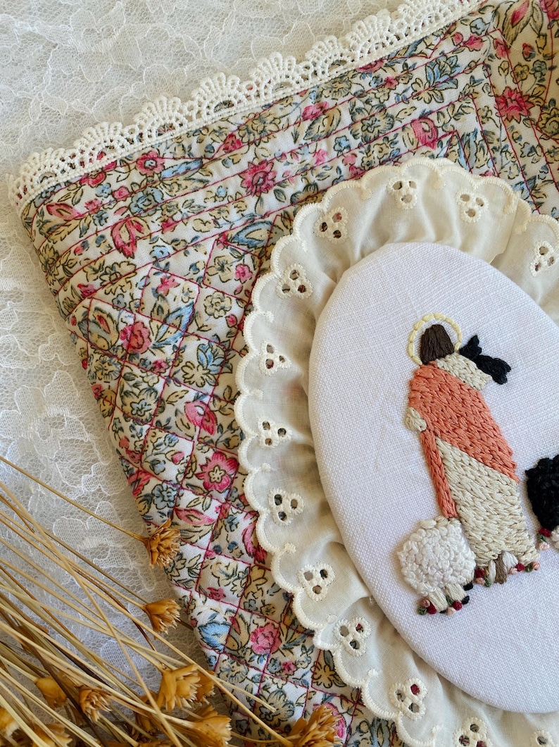 May include: An oval embroidery hoop featuring a stitched figure in a peach robe with a halo, standing beside a white sheep. The hoop is framed by a white eyelet lace trim and rests on a quilted fabric with a floral pattern and lace edge.