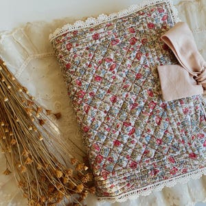 May include: A quilted fabric book cover featuring a floral pattern in shades of red, blue, and green. The cover is edged with white lace trim and includes a light pink fabric bow attached to the front. The design has a diamond-stitched texture.