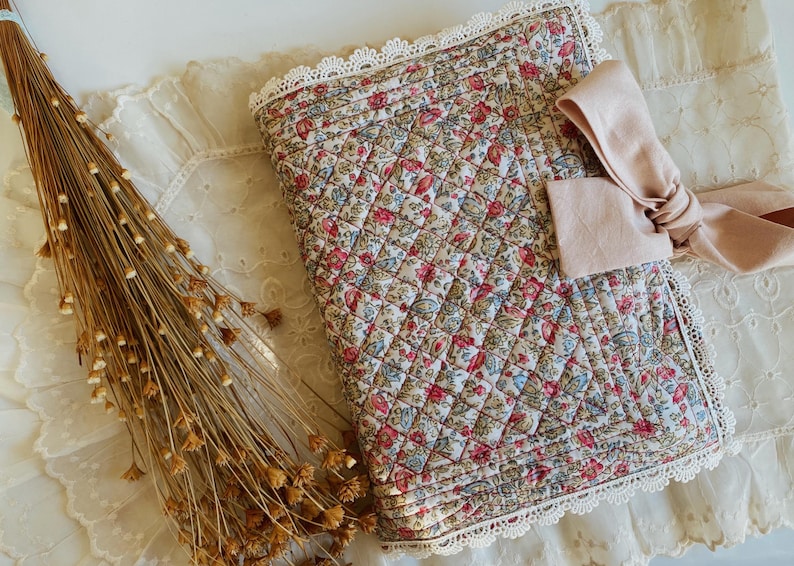 May include: A quilted fabric book cover featuring a floral pattern in shades of red, blue, and green. The cover is edged with white lace trim and includes a light pink fabric bow attached to the front. The design has a diamond-stitched texture.