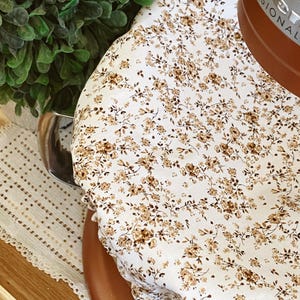 May include: A white floral patterned cover with brown flowers and green leaves. The cover is draped over a brown object. A green plant and a decorative white lace mat are in the background.