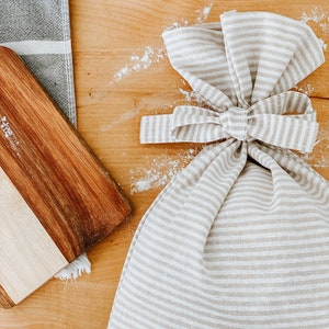 May include: A beige and white striped linen flour sack with a bow tied at the top. The sack is sitting on a wooden surface with a wooden cutting board and a gray and white striped kitchen towel.