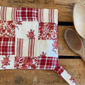 May include: Red and white patchwork pot holder with a floral design. The pot holder has a loop for hanging.
