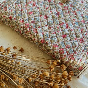 May include: A quilted fabric pouch featuring a floral pattern with pink, blue, and green blossoms on a cream background. The rectangular pouch is finished with white lace trim along the edges and a light pink fabric bow attached to the top corner.