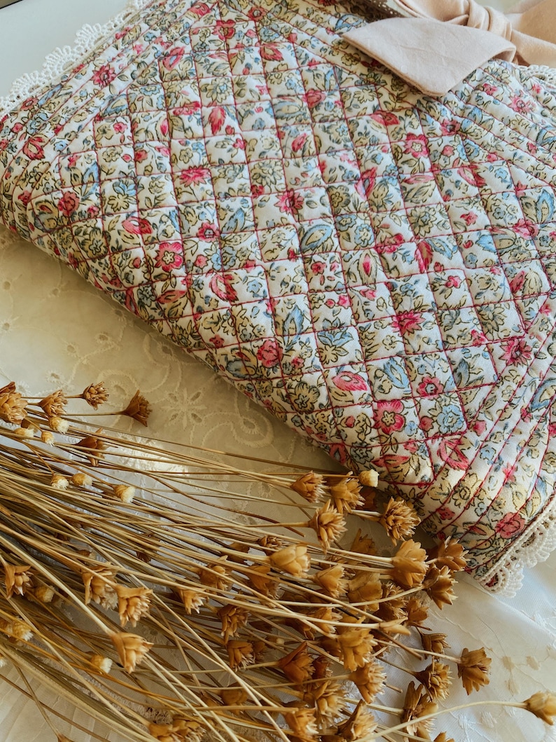 May include: A quilted fabric pouch featuring a floral pattern with pink, blue, and green blossoms on a cream background. The rectangular pouch is finished with white lace trim along the edges and a light pink fabric bow attached to the top corner.