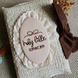 May include: A light green and white patterned book cover with an embroidered oval patch. The patch reads "holy bible" and "Romans 8:39" in brown thread. A white lace trim surrounds the patch. A brown ribbon and dried flowers are also visible.
