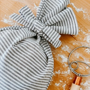 May include: A white and gray striped cloth bag tied with a bow, sitting on a wooden surface with white flour scattered around it. A copper and wooden dough hook is also visible in the image.
