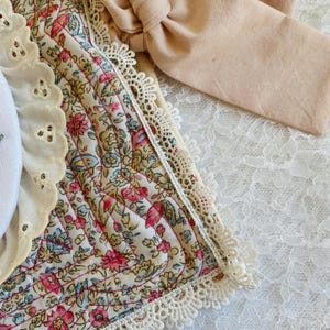 May include: A quilted fabric piece featuring a floral pattern in pink, blue, and green on a cream background. It is trimmed with white scalloped lace and includes a solid beige fabric tie. A white eyelet lace border accents the side.