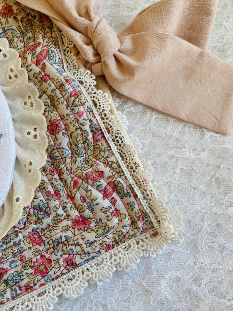 May include: A quilted fabric piece featuring a floral pattern in pink, blue, and green on a cream background. It is trimmed with white scalloped lace and includes a solid beige fabric tie. A white eyelet lace border accents the side.
