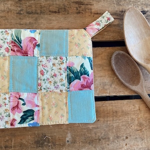 May include: A patchwork pot holder with a floral pattern in pink, yellow, and blue. The pot holder has a loop for hanging. Two wooden spoons are next to the pot holder on a wooden surface.