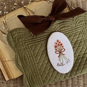 May include: An olive green quilted placemat with a brown bow and an embroidered oval. The oval features a bouquet of pink flowers tied with a brown ribbon and the text "Ruth 1:17". An open book and floral garland are also present.