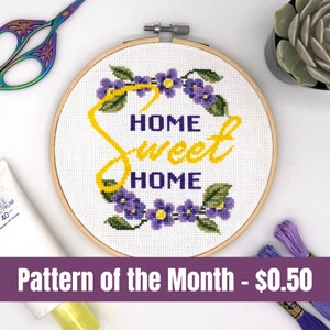 May include: A cross-stitch pattern featuring a wreath of purple flowers with green leaves surrounding the text "Home Sweet Home" in yellow lettering. The pattern is displayed in a wooden embroidery hoop.