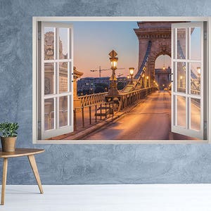 Puede incluir: Una vista de ventana de un puente con farolas y una ciudad en el fondo. El puente está hecho de piedra y tiene una barandilla de metal. Las farolas están encendidas y proyectan un brillo cálido sobre el puente. La ciudad está en la distancia y está iluminada por el sol poniente.