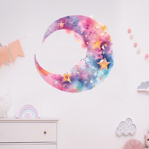 Peut inclure: Un autocollant mural en forme de croissant de lune en aquarelle avec un fond rose, bleu et violet. La lune est remplie d'étoiles dorées et de volutes.