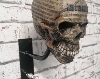 May include: A decorative skull made of resin with a printed design of text and the word "Dragons" on the forehead. The skull is mounted on a black metal wall bracket.