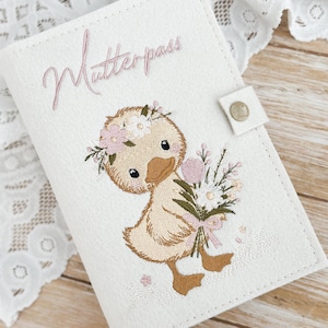 Könnte beinhalten: Eine weiße, rechteckige Dokumentenhülle mit einem gestickten hellbraunen Entenküken, das einen Blumenkranz trägt und einen Blumenstrauß hält. Der Schriftzug "Mutterpass" ist in rosa Schreibschrift über dem Entenküken gestickt. Mit Druckknopfverschluss.