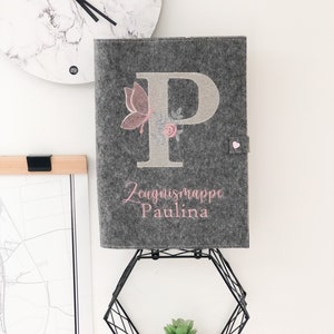 Könnte beinhalten: Grauer Filzordner mit einem rosa Schmetterling und einer Blumenstickerei. Der Ordner hat einen großen gestickten Buchstaben "P" und den Text "Zeugnismappe Paulina".