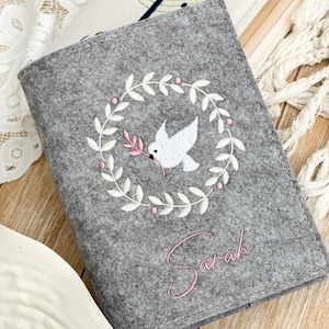 May include: Gray felt book cover with white embroidery of a dove with an olive branch in its beak inside a wreath. The name "Sarah" is embroidered in pink below the wreath.