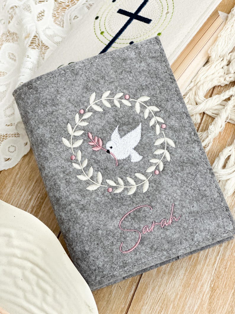 May include: Gray felt book cover with white embroidery of a dove with an olive branch in its beak inside a wreath. The name "Sarah" is embroidered in pink below the wreath.