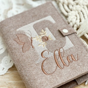 Könnte beinhalten: Ein taupefarbener Filz-Reisepasshalter mit dem Namen "Ella" in Kupfer gestickt. Der Buchstabe "E" ist cremefarben und mit einem Schmetterling- und Blumendesign bestickt. Er hat einen Druckknopfverschluss.