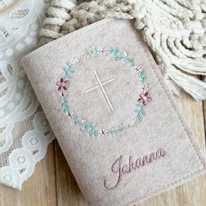 May include: A beige felt book cover with a white cross embroidered in a circle of pink and green flowers. The name "Johanna" is embroidered in pink below the cross.