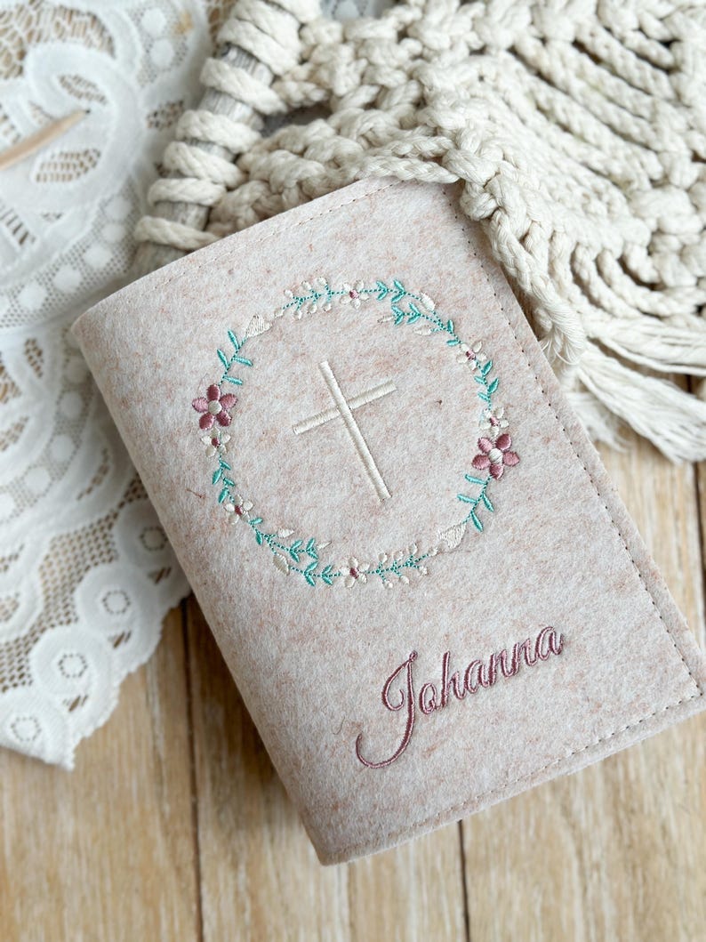 May include: A beige felt book cover with a white cross embroidered in a circle of pink and green flowers. The name "Johanna" is embroidered in pink below the cross.