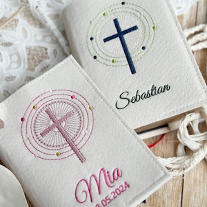 May include: Two white felt book covers with embroidered crosses and names. The top cover has a blue cross with a green circle around it and the name "Sebastian". The bottom cover has a pink cross with a pink circle around it and the name "Mia" with the date "18.05.2024" below.