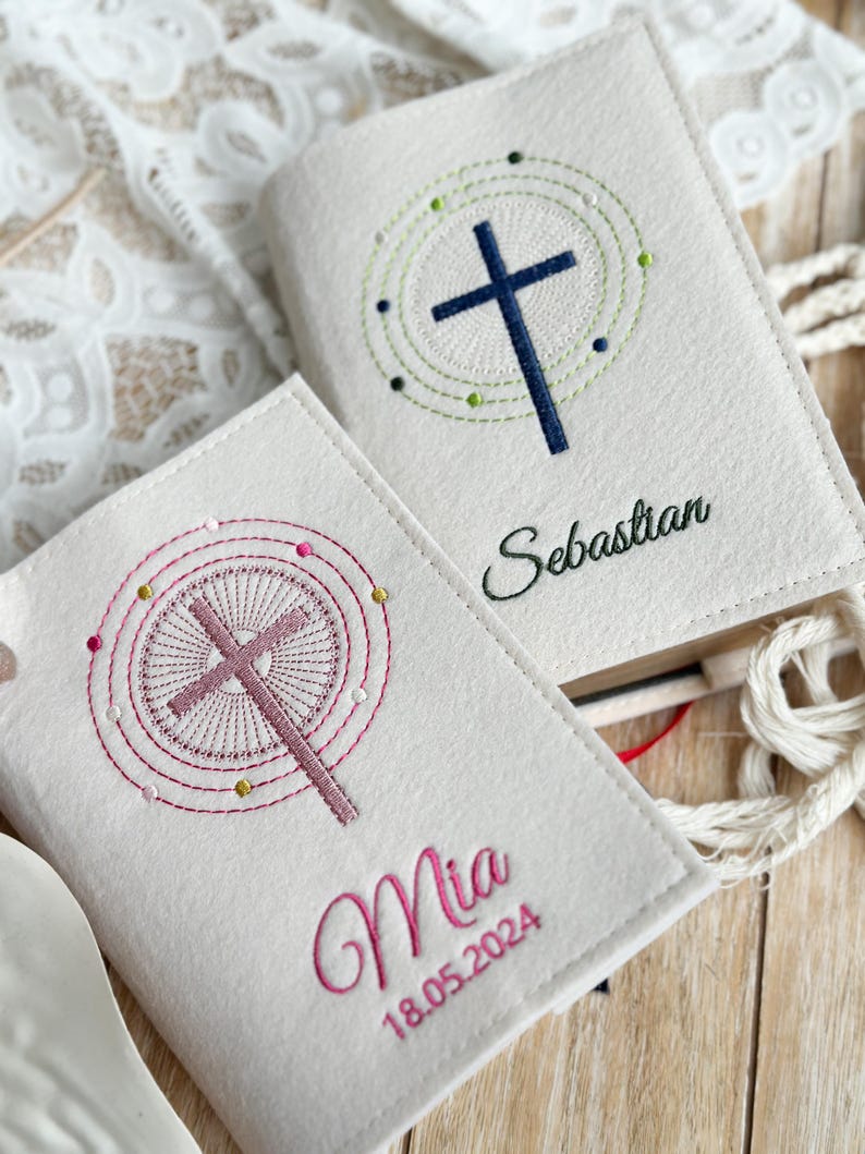 May include: Two white felt book covers with embroidered crosses and names. The top cover has a blue cross with a green circle around it and the name "Sebastian". The bottom cover has a pink cross with a pink circle around it and the name "Mia" with the date "18.05.2024" below.