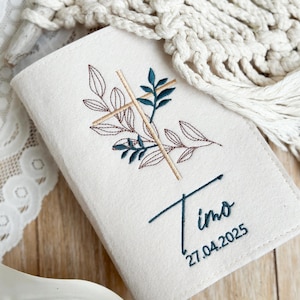 May include: A white felt book cover with a cross and leaves embroidered in brown and green thread. The name "Timo" and the date "27.04.2025" are also embroidered in green thread.