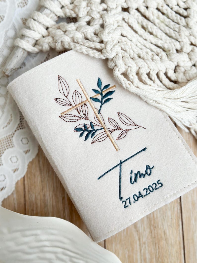 May include: A white felt book cover with a cross and leaves embroidered in brown and green thread. The name "Timo" and the date "27.04.2025" are also embroidered in green thread.