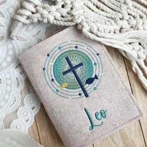 May include: A beige felt book cover with an embroidered design of a cross, fish, and circles in shades of green, blue, and yellow. The name "Leo" is embroidered in green below the design.