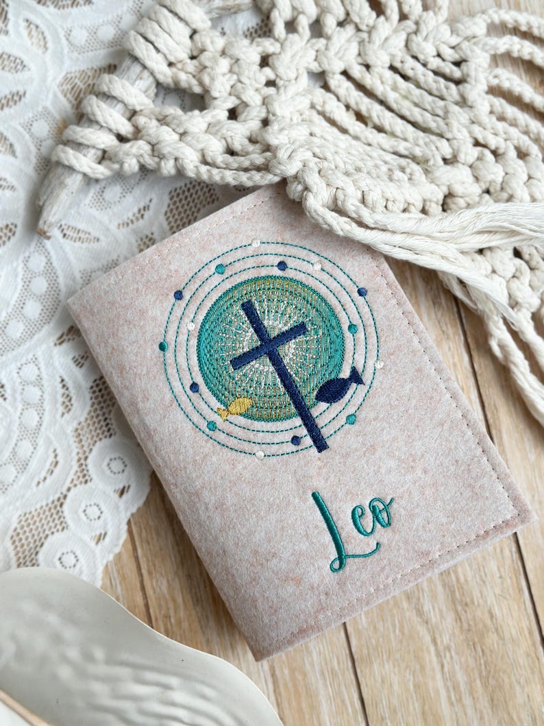 May include: A beige felt book cover with an embroidered design of a cross, fish, and circles in shades of green, blue, and yellow. The name "Leo" is embroidered in green below the design.