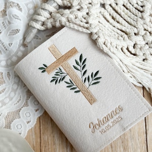 May include: A white felt book cover with an embroidered cross and green leaves. The text "Johannes 10.05.2025" is embroidered below the cross.